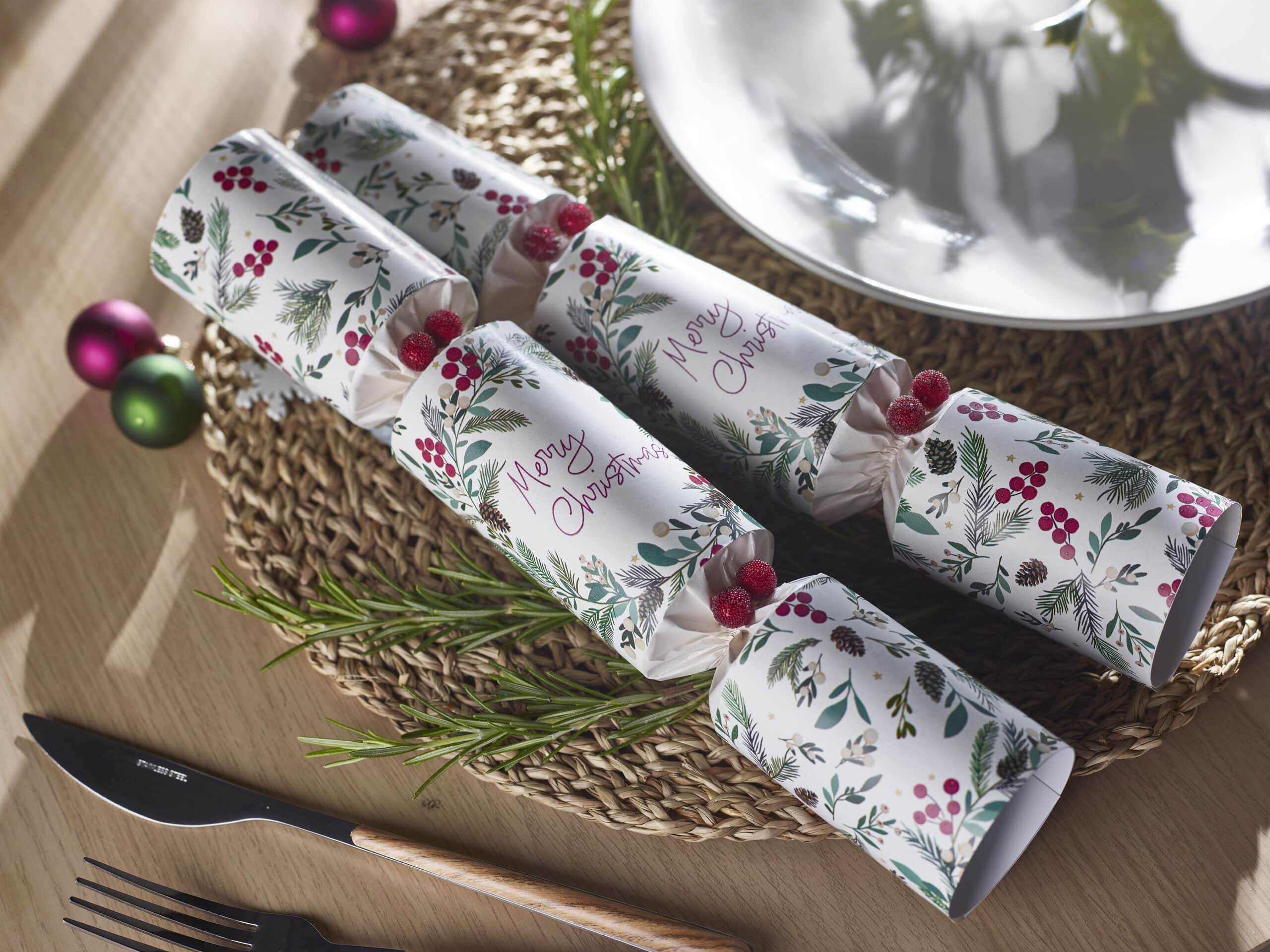Christmas crackers on a woven placemat with rosemary sprigs, red berries, and a knife and forks on a wooden table, festive scene.