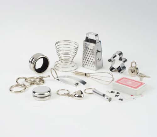Assorted stainless steel kitchen tools scattered on a white surface, including a whisk, grater, rings, and a deck of playing cards nearby.