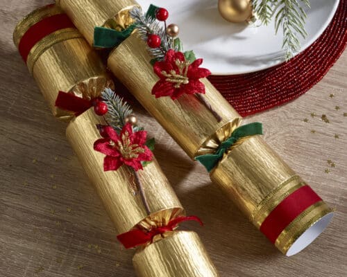 Gold-wrapped gifts with red ribbons, poinsettia accents, pine sprigs, and a red beaded garland on a wooden table for Christmas decor.